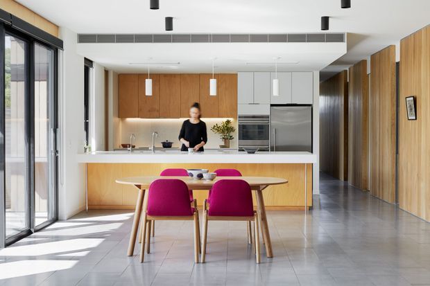 Bright open-plan kitchen and dining area with a streamlined contemporary fit-out. The kitchen features extensive warm timber cabinetry, a white island bench, integrated stainless-steel appliances, and a dining table with vivid magenta upholstered chairs. Full-height black-framed glazing brings in strong natural light, while grey tiled flooring and a timber-lined hallway complete the space.