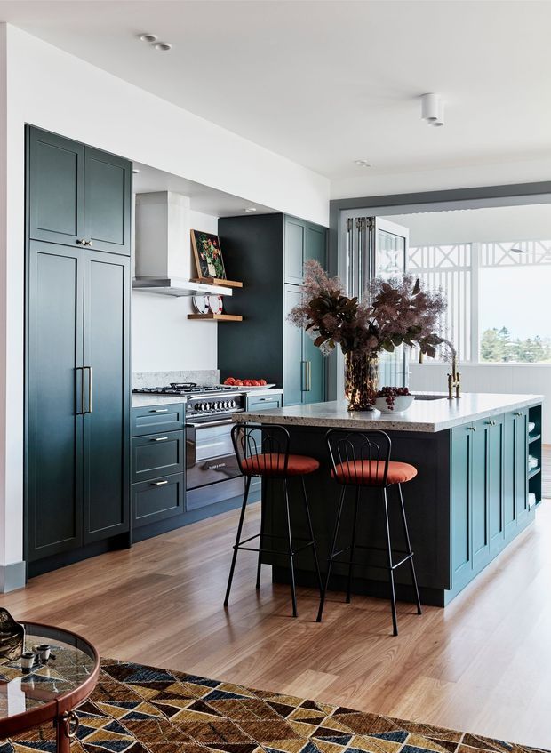 A refined contemporary kitchen with deep blue-green paneled cabinetry, a large island with a pale stone benchtop, integrated seating, a freestanding stainless-steel range, and open shelving. Warm timber flooring, black metal bar stools with red upholstered seats, and a patterned rug add contrast. Large glazing brings in abundant daylight and views beyond.