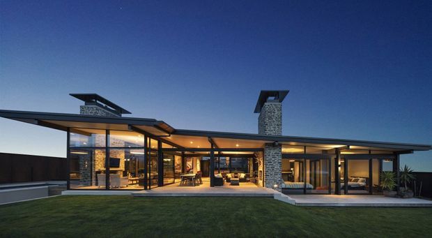 Twilight exterior view of a low, single-storey modern home with extensive floor-to-ceiling glazing, broad projecting roof planes, dark slender frames, and prominent stone-clad chimney volumes. Warm interior lighting reveals open-plan living areas, while a lawn and paved terrace extend across the foreground.