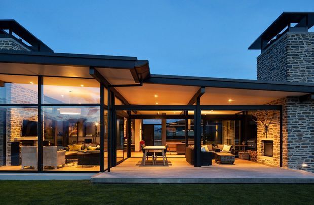 Exterior dusk view of a contemporary single-storey residence with extensive floor-to-ceiling glazing, dark metal framing, low-pitched flat roof forms, and prominent stacked natural-stone masonry walls. The open glazed central section reveals a dining and living area, while a covered timber-toned deck extends across the front. Warm interior and recessed exterior lighting create an inviting contrast against the blue evening sky.
