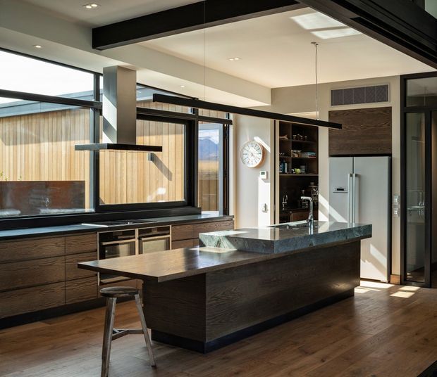 Contemporary kitchen with a large dark timber island, integrated dining overhang, dark stone benchtop, extensive black-framed glazing, and timber cabinetry. A stainless-steel refrigerator and open shelving sit against the rear wall, while linear pendant lights and concealed ceiling lighting illuminate the space. Warm timber flooring contrasts with the black and charcoal joinery, and the glazing looks toward a timber-clad exterior courtyard or boundary.