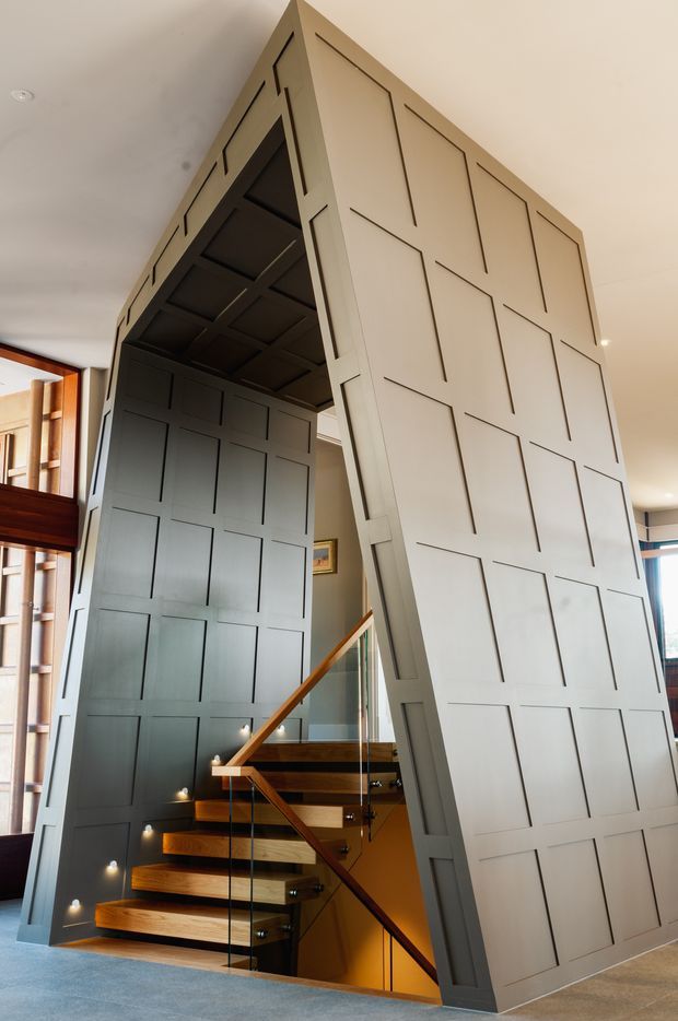 Interior view of a dramatic stairwell enclosed by tall, angled wall panels with recessed rectangular detailing. The staircase has warm timber treads and handrails, clear glass balustrades with dark fittings, integrated step lighting, and a dark timber-paneled underside. Neutral painted walls and carpeted flooring surround the sculptural stair volume, with timber-framed glazing visible at the side.