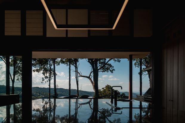 Dark, refined interior opening onto a reflective infinity-edge pool and expansive floor-to-ceiling glazing, with a wooded and coastal-looking landscape beyond. The room features a glossy dark island or counter, integrated black tapware, timber wall panelling, and a dramatic linear recessed ceiling light.