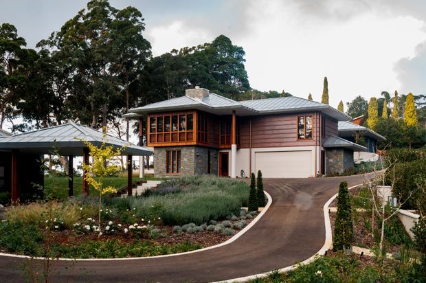 Exterior view of a substantial contemporary home set within a landscaped garden. The house combines horizontal timber-clad upper walls, dark stone masonry, extensive timber-framed glazing, and multiple low-pitched metal hipped roof forms. A curved driveway leads to an integrated double garage, with a separate open-sided carport structure visible to the left.
