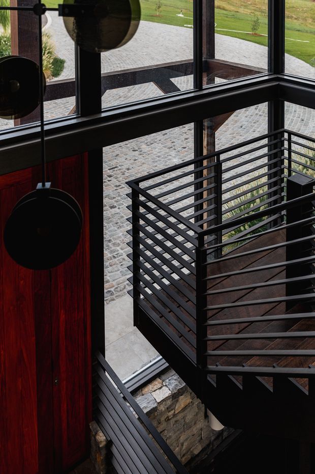 A dramatic interior view of a modern stairwell with dark steel horizontal railings, a timber stair flight, expansive black-framed glazing, and a dark red timber wall panel. Natural stonework forms the lower stairwell wall, while a paved courtyard and landscaped grounds are visible outside.