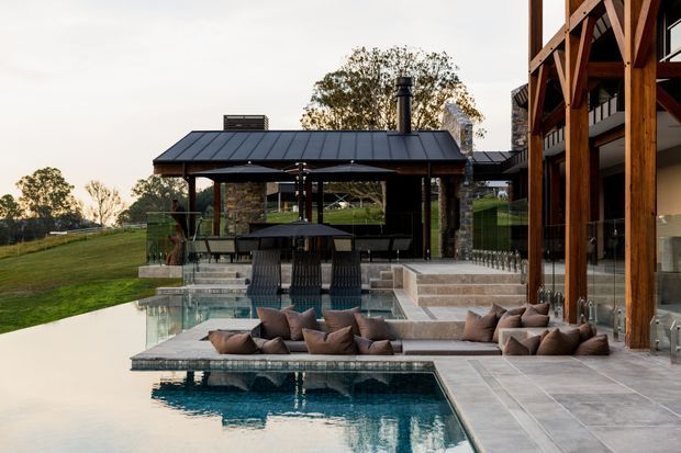 A sophisticated outdoor entertaining area beside a modern residence, featuring a large reflective swimming pool, sunken lounge seating, a covered pavilion with dining furniture, and a dark metal roof. The adjoining house has extensive glazing, substantial timber posts and beams, and stone-clad elements. The landscaped rural setting is visible beyond the pool.