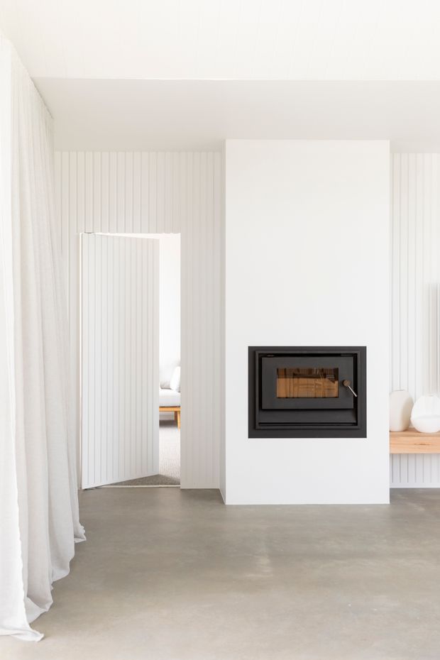 Bright minimalist interior featuring a white plaster fireplace surround with a built-in black wood-burning fireplace, vertical white wall panelling, pale curtains, a smooth grey concrete floor, and a light timber built-in bench with simple white objects. An open doorway leads to an adjacent room.
