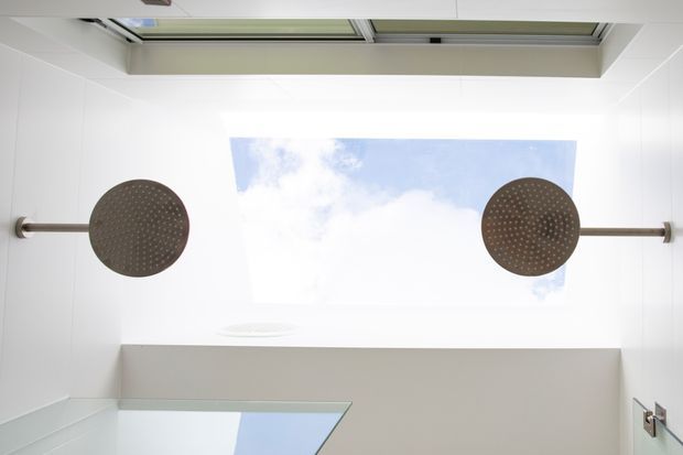 Upward view inside a bright, minimalist bathroom showing a large central skylight with blue sky visible, two round wall-mounted rainfall showerheads on horizontal metal arms, white wall and ceiling surfaces, and a partial glass shower enclosure.