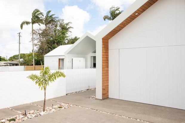 Exterior view of a bright, minimalist residential property with white rendered walls, a large white garage door, and a sharply angled gable-like roof edge lined with warm timber soffit and trim. A paved driveway and low white boundary wall frame the foreground, with tropical planting visible around the home.