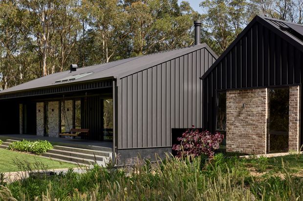 Exterior view of a contemporary single-storey residential building composed of connected pitched-roof volumes. The facade combines dark vertical cladding, a dark metal roof, and exposed warm-toned brick around a recessed opening. A covered entry or veranda with steps, glazing, and landscaping fronts the building, with mature trees in the background.