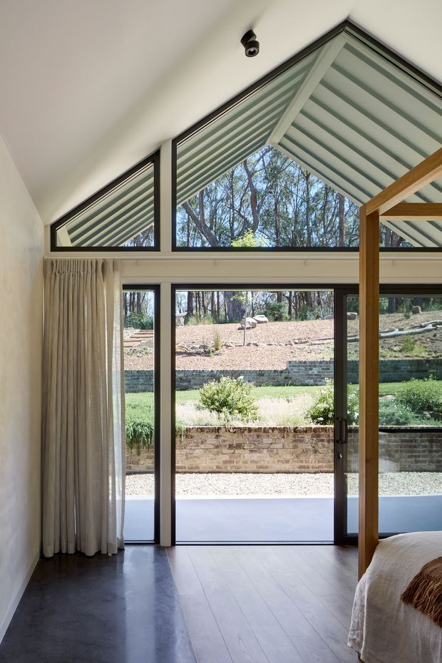 A bright bedroom interior with a timber four-poster bed partially visible at right, opening onto a landscaped outdoor area through large dark-framed glazed doors and triangular clerestory windows. The room has a high sloping ceiling, sheer full-height curtains, smooth white walls, and contrasting dark polished flooring beside pale timber floorboards. The glazing reflects or reveals a striped exterior shading structure and surrounding trees.