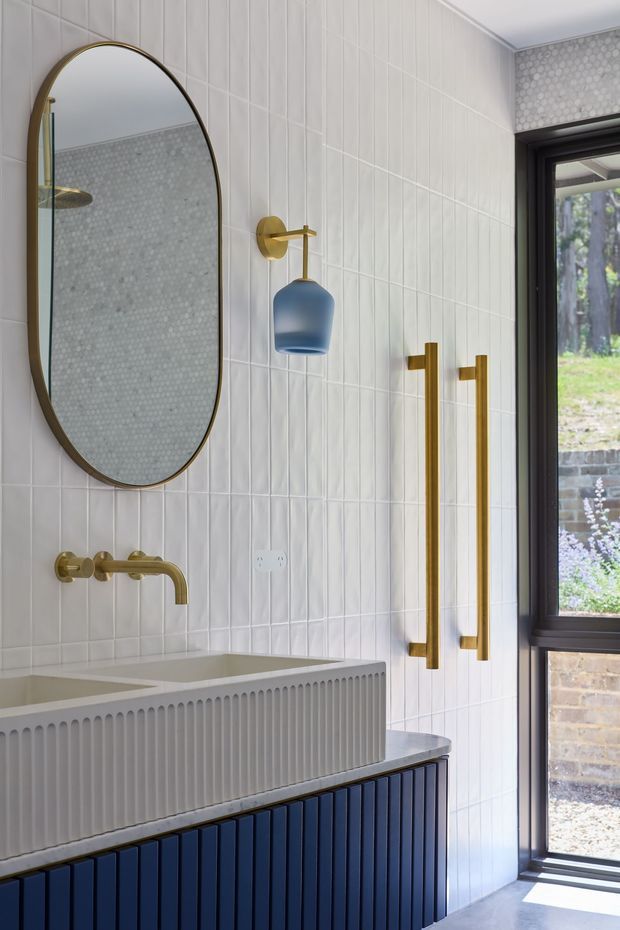 A refined contemporary bathroom vanity area with a large arched brass-framed mirror, white vertically fluted wall tiles, a white ribbed basin, dark navy-blue fluted vanity cabinetry, and brushed brass wall-mounted tapware and hardware. A blue pendant light hangs beside the mirror, while a black-framed glazed door or window provides views to a landscaped exterior.
