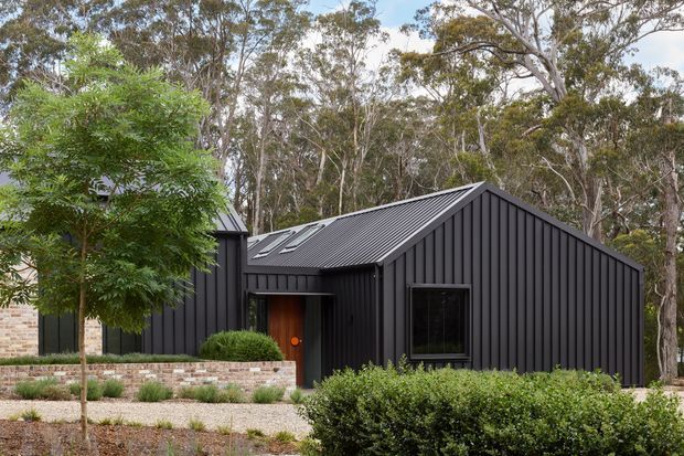 Exterior view of a contemporary single-storey home set within a eucalyptus woodland landscape. The building has dark vertical metal cladding, a steep gabled roof with matching dark corrugated roofing, black-framed windows, and a recessed entry with a warm timber door. Low native-style planting, gravel landscaping, and a low mixed brick-and-stone retaining wall frame the facade.