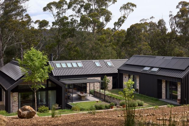 A low-rise modern residential complex set within a wooded landscape, composed of several connected or closely grouped dark-clad pavilion-like volumes. The buildings have steeply pitched metal roofs with multiple skylights, extensive glazing, and sections of exposed light brick masonry. A landscaped courtyard and covered outdoor area sit between the wings, with a gravelly sloped foreground and dense eucalyptus-like bushland behind.