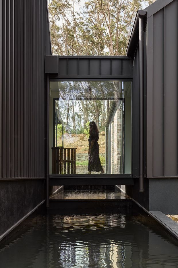 A dramatic exterior architectural view of a narrow water channel running between two dark, vertically clad building volumes. A glazed bridge or passage spans the channel, reflecting surrounding trees and revealing a figure inside. The composition highlights black metal framing, corrugated/ribbed wall cladding, exposed structural elements, and strong indoor-outdoor connection in a wooded setting.