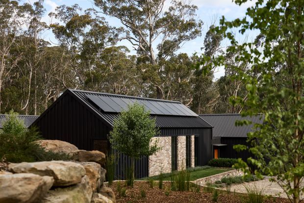 A dark, low-profile gabled house set among mature eucalyptus woodland. The exterior combines black vertically ribbed cladding and dark metal roofing with pale textured brick masonry around the entrance. A landscaped foreground includes large natural stone retaining elements, gravel paving, lawn, and native-style planting. The composition presents the building exterior within a wooded Australian-like setting.