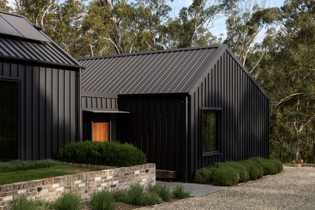 Exterior view of a compact contemporary building composed of dark vertical metal-clad volumes with steep gable roofs and matching dark metal roofing. A warm timber entry door sits beneath a small projecting canopy, with low landscaped planting, a brick retaining wall, concrete path, and gravel foreground. The building is set among mature eucalyptus trees.