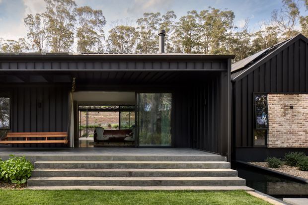 Exterior view of a contemporary dark-clad home set among eucalyptus trees. The single-storey composition combines a flat-roofed pavilion with a steep gabled volume, broad glazed openings, a covered entry terrace, and wide concrete steps. A brick feature wall is visible within the gabled section, while a bench and interior furnishings can be seen through the glazing.