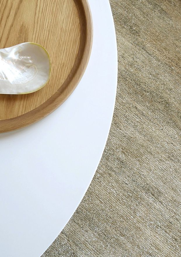 Close-up detail of a minimalist interior vignette showing a white curved tabletop, a round light-oak serving tray, and a small mother-of-pearl dish, positioned over a neutral beige textured carpet.
