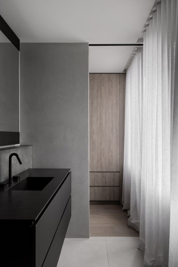 A refined, minimalist interior view showing a dark stone kitchen island with integrated black sink and curved tap in the foreground. A tall smooth grey partition separates the kitchen from a warm light-wood built-in storage area, while full-height sheer curtains line the right side and diffuse natural light. The palette is monochromatic grey, black, white, and light timber, with clean linear detailing and concealed cabinetry.