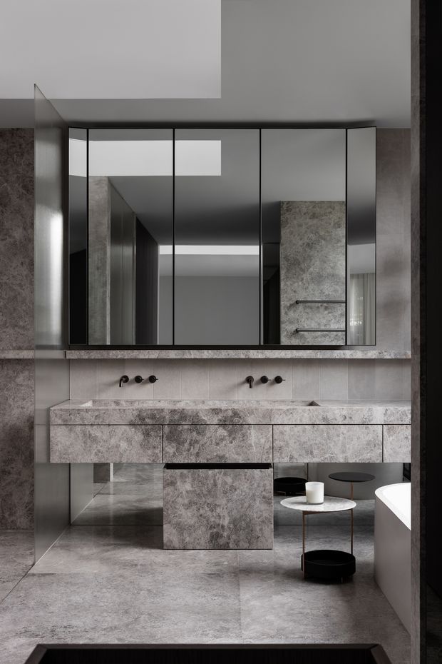 A sophisticated monochromatic bathroom with a long double vanity clad in veined grey stone, two wall-mounted black tap sets, large mirrored cabinetry above, and a freestanding white bathtub at the right. The room features large grey stone-look floor and wall surfaces, black-framed details, integrated storage, and small black side tables and stools. The styling is highly minimal, refined, and contemporary with strong geometric lines and reflective surfaces.