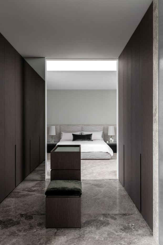A refined, dark-toned dressing corridor or walk-in wardrobe leading directly into a minimalist bedroom. Floor-to-ceiling dark timber cabinetry lines both sides, with a central built-in dark timber island and upholstered bench. Large-format grey stone-look flooring continues toward the bedroom, which features a low upholstered bed, white bedding, bedside lamps, and a pale upholstered headboard. The space has a restrained, luxurious contemporary feel with a bright ceiling opening or clerestory above the bedroom entrance.