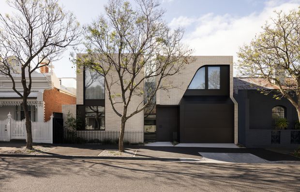 Street-facing contemporary multi-storey residence with a restrained dark and warm-neutral facade. The building combines pale textured masonry, dark metal-look cladding, large glazed openings, and a strongly framed curved upper window, set among mature street trees and adjacent traditional homes.