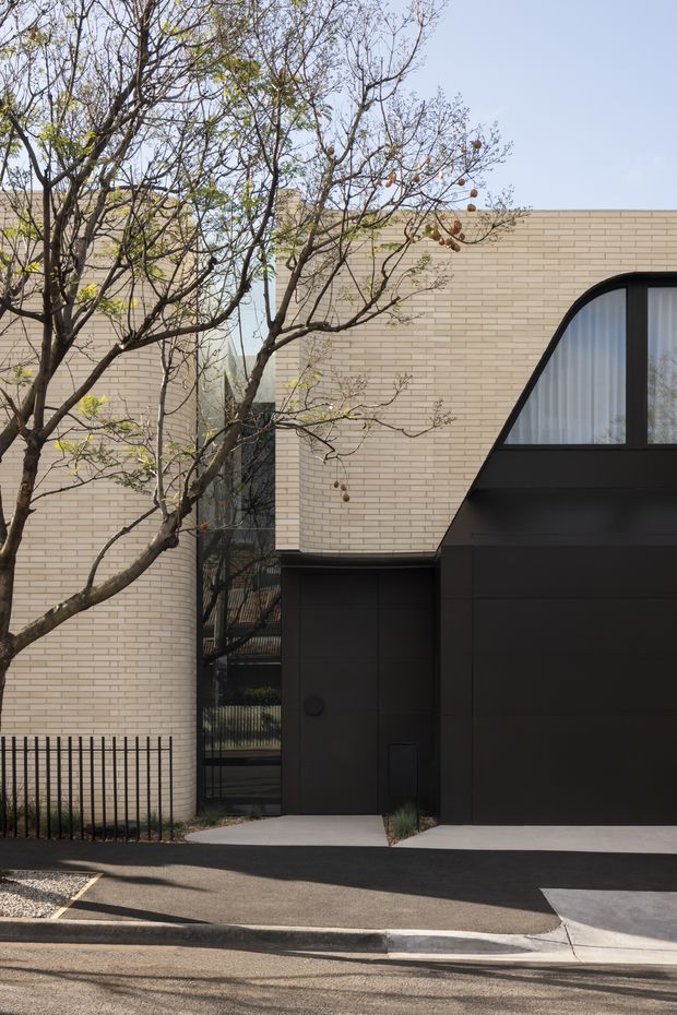Street-facing view of a refined contemporary residential facade, combining pale cream brickwork with a dark charcoal metal-clad entry and garage volume. A distinctive large arched window with sheer curtains punctuates the upper facade, while mature deciduous trees and a slim black metal fence frame the frontage.