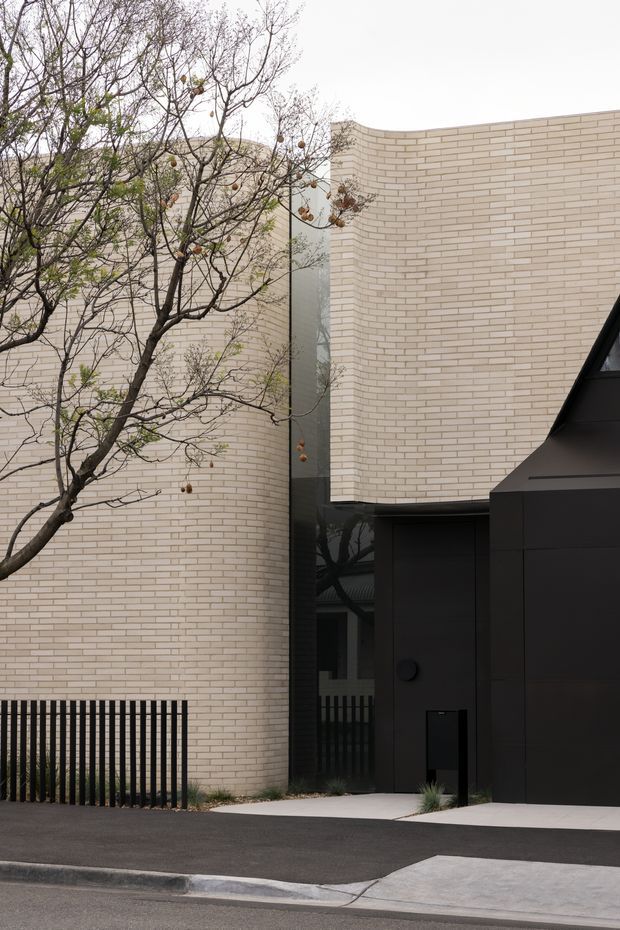 A contemporary residential facade viewed from the street, featuring pale cream brickwork with curved wall geometry, a tall narrow dark glazed opening, and a bold matte-black entry or garage volume. A simple black vertical-bar fence, concrete footpaths, and sparse deciduous planting complete the restrained streetscape.