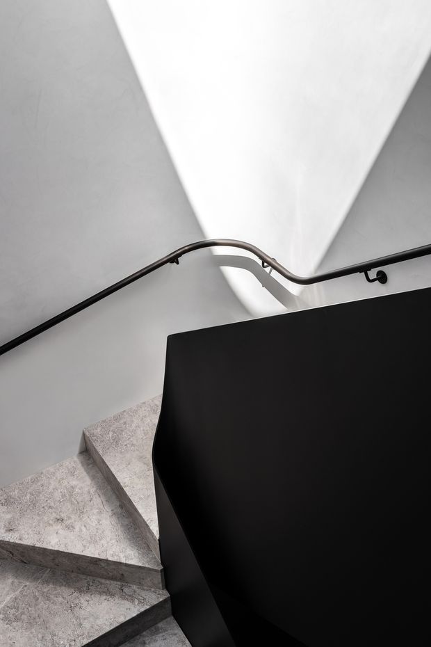 A close-up architectural view looking down a modern stairway. Light grey stone or concrete treads turn around a large matte-black central balustrade or stair enclosure, with slim dark metal handrails fixed to white plaster walls. Strong geometric shadows and a narrow skylight or roof opening create a dramatic monochrome composition.