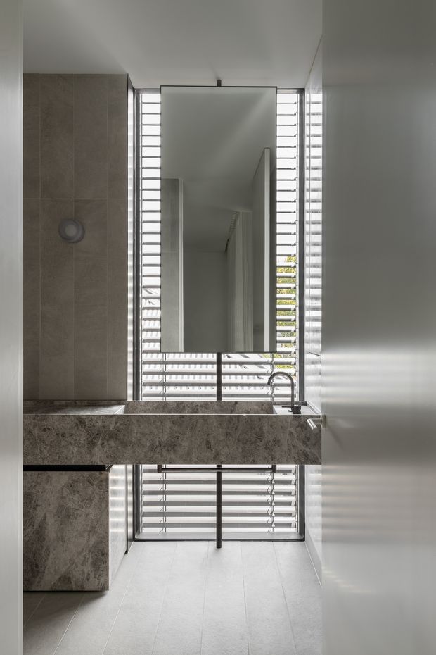 A narrow, contemporary bathroom viewed toward a large wall mirror above a stone vanity. The room features grey stone-look wall and vanity surfaces, pale tiled flooring, a minimalist basin with dark metal tapware, and full-height louvred windows bringing in filtered daylight.