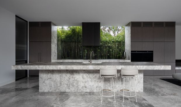 Contemporary kitchen with a large grey stone island and waterfall-style stone base, integrated sink and tap, dark timber cabinetry, and two light upholstered bar stools. Full-height glazing behind the island looks onto dense greenery, while dark stone or concrete-look flooring and restrained finishes create a refined, monochromatic interior.