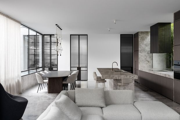 Open-plan contemporary living, dining and kitchen interior with a neutral monochromatic palette. A large light-grey sectional sofa occupies the foreground, with a dark dining table and upholstered chairs behind it. The kitchen features a stone island with integrated sink, dark cabinetry, full-height stone-look wall finish and black-framed glazed doors. Floor-to-ceiling glazing with sheer curtains and a sculptural pendant add an airy, refined feel.