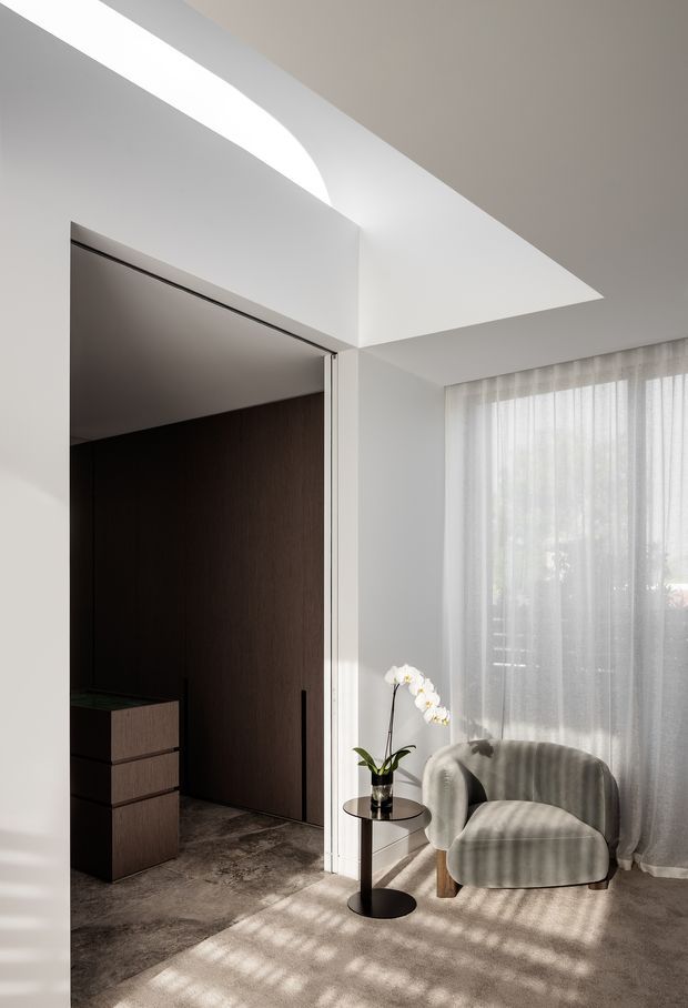 A refined contemporary interior corner with a sculptural curved armchair beside a small round black side table and orchid. Sheer full-height curtains filter daylight into the room, creating patterned shadows across a neutral carpet. An open doorway reveals a dark timber-lined adjoining space with built-in cabinetry, while a crisp white ceiling features a geometric recessed or angled detail.