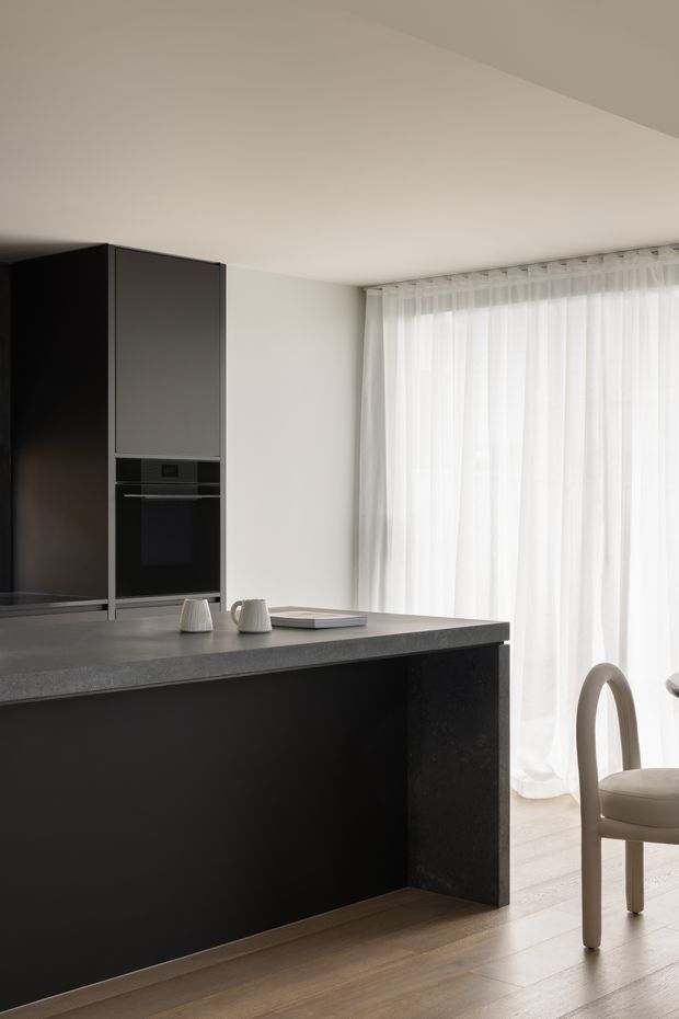 A refined contemporary kitchen interior featuring a large dark island with a grey stone benchtop, integrated dark cabinetry and appliances, pale timber flooring, and sheer white curtains across a bright window. A minimal dining chair and small neutral accessories are visible beside the island.