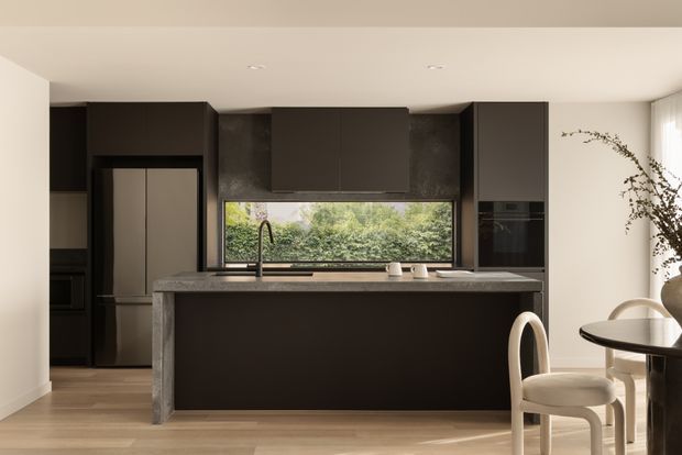 A contemporary kitchen with dark full-height cabinetry, a large central island, integrated appliances, and a stone-look benchtop. A horizontal window above the sink overlooks greenery, while light timber flooring and warm neutral walls balance the dark joinery. A small adjoining dining area with a round table and upholstered chairs is visible at right.