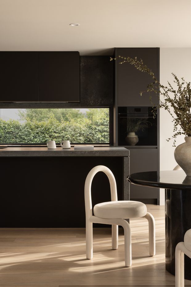 A refined contemporary kitchen and dining area with matte black full-height cabinetry, a dark stone benchtop, integrated appliances, and a large horizontal window overlooking dense greenery. A sculptural cream dining chair and dark round dining table sit in the foreground, with warm timber flooring, neutral walls, and soft natural light creating a sophisticated, high-contrast interior.