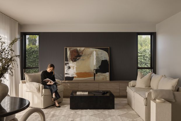 A refined living room with a large cream sofa and armchair arranged around a dark rectangular coffee table. The room features warm timber built-in cabinetry, a dark vertical fluted feature wall, black-framed windows, soft neutral furnishings, and a large abstract artwork. Natural light and greenery are visible through the windows.