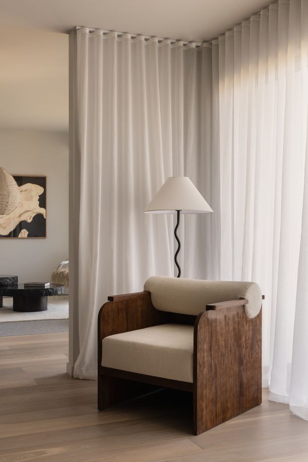 A softly lit contemporary living room corner featuring a sculptural wood-framed armchair with cream upholstery, a minimalist floor lamp, full-height sheer white curtains, and warm timber flooring. A coffee table, artwork, and additional seating are partially visible in the adjoining space.