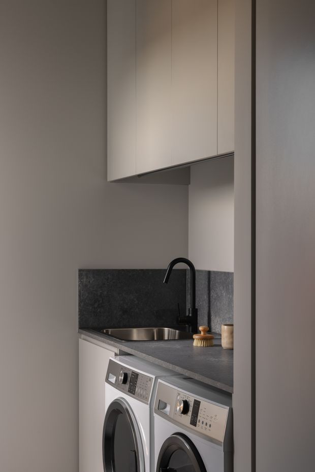Compact, contemporary laundry room with stacked? side-by-side front-loading washing machine and dryer beneath a dark stone benchtop. The space features a stainless-steel undermount sink, matte black gooseneck tap, dark stone splashback, and tall light-grey built-in overhead cabinetry. Neutral grey finishes and integrated storage create a clean, minimalist look.