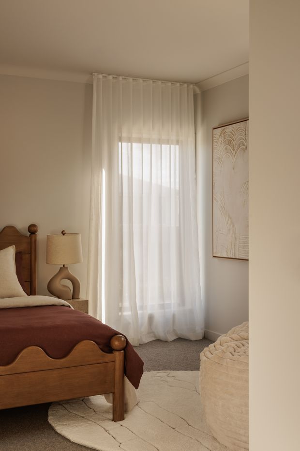 A warm, softly lit bedroom with a traditional wooden bed featuring turned posts and a scalloped footboard. The room has sheer full-height curtains over a window, beige walls with crown moulding, a neutral textured rug, a sculptural bedside lamp, framed abstract artwork, and a rounded upholstered ottoman or bench.