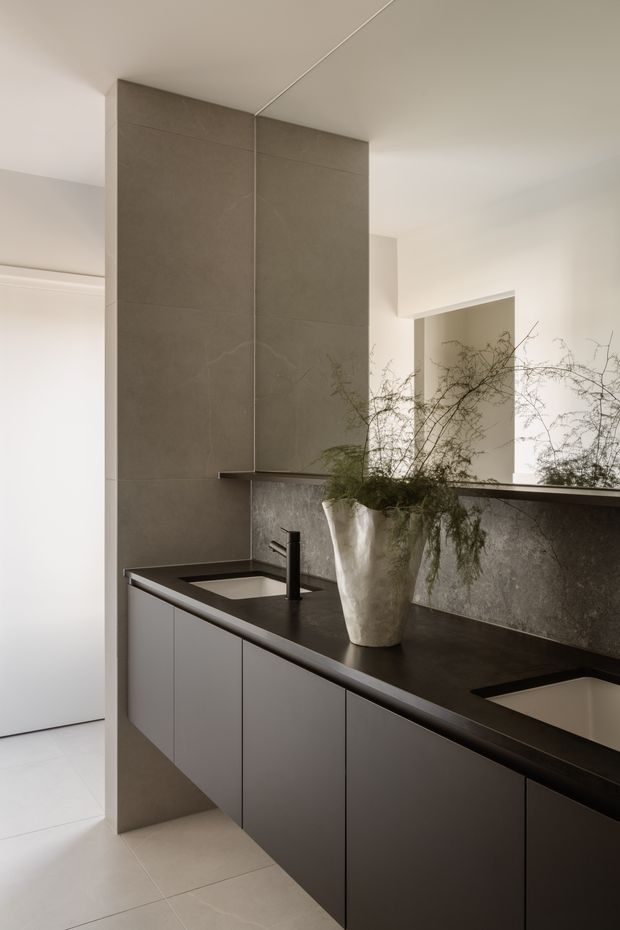 A refined minimalist kitchen or kitchenette with dark matte cabinetry, a black stone benchtop, integrated sinks and tapware, and a tall grey stone-clad wall. A large mirror reflects the counter and sparse branch arrangement, while pale tiled flooring and off-white walls create a calm, understated interior.
