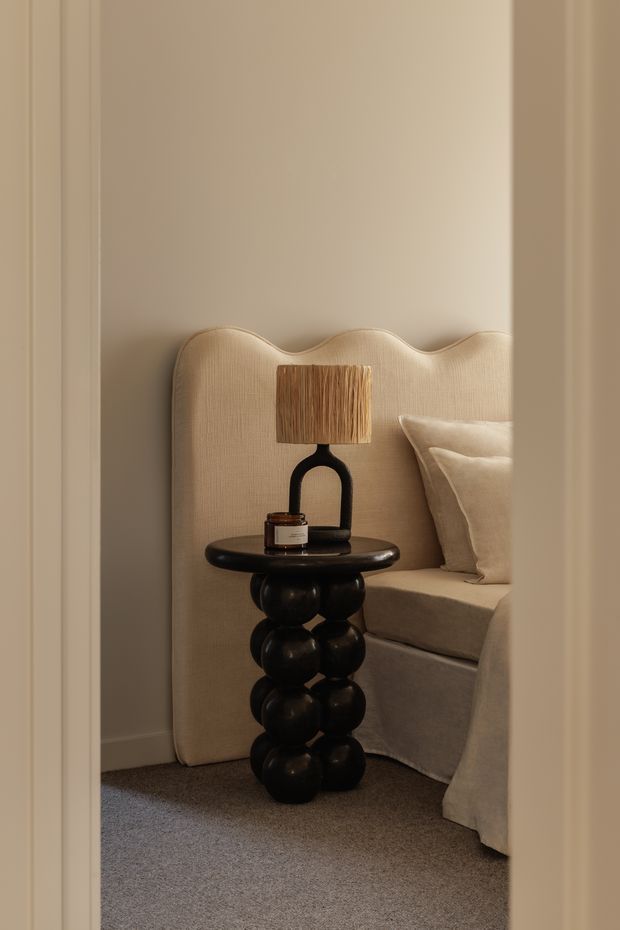 A softly lit bedroom vignette viewed through a doorway, featuring a sculptural upholstered headboard in warm beige, layered neutral bedding, grey-beige carpet, and a glossy black rounded bedside table with a matching black loop-base lamp and textured shade. The restrained palette and curved forms create an intimate, refined contemporary interior.