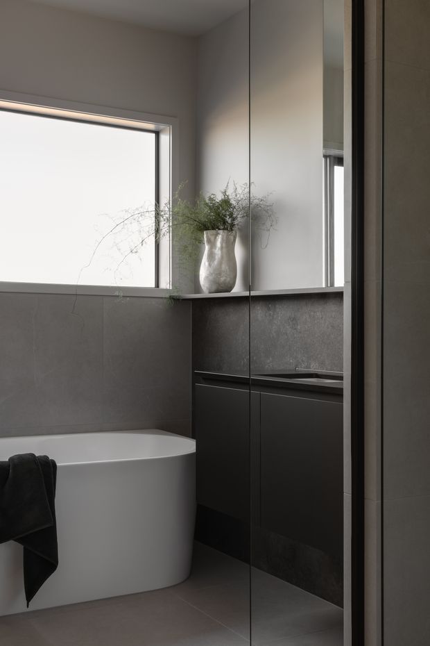 A refined contemporary bathroom with a freestanding white bathtub, dark charcoal wall and floor tiles, and a dark floating vanity with an integrated basin. A large frameless mirrored cabinet reflects the vanity and window, while a bright rectangular window and a sculptural vase with delicate greenery add natural light and softness.