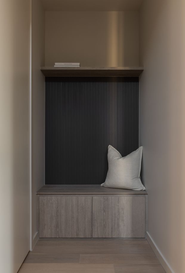 A narrow built-in alcove featuring a dark vertically fluted wall panel, a timber-toned shelf and bench seat, and integrated wood-grain cabinetry below. A single light neutral cushion rests on the bench, while warm taupe walls and timber flooring create a restrained, refined interior.