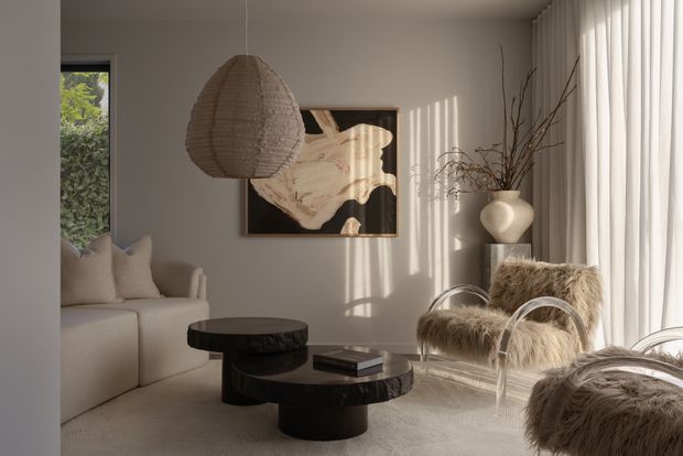 Warm, softly lit contemporary living room with a neutral palette. The space features a large cream sectional sofa, layered dark rounded coffee tables, sculptural furry lounge chairs, full-height sheer curtains, a woven pendant light, and abstract wall art. Sunlight creates strong vertical shadows across the walls and furniture, giving the room a refined, cozy atmosphere.
