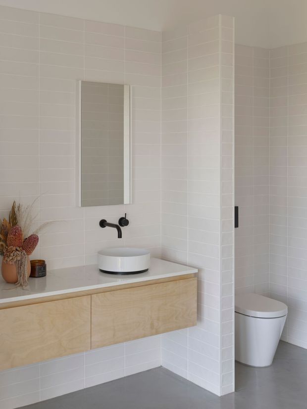 Bright contemporary bathroom with white rectangular wall tiles, a floating pale timber vanity, white vessel basin, matte black wall-mounted tapware, frameless mirror, and a partially screened toilet. The space has a clean, minimal finish with warm natural accents from the vanity and dried decorative arrangement.