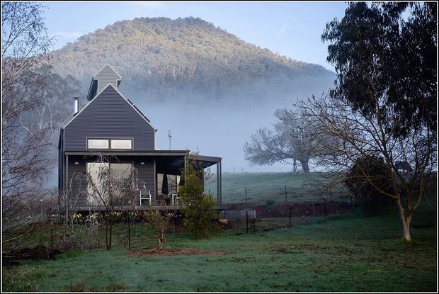 A dark-clad rural house with a steep gabled roof, projecting upper dormer, and covered veranda sits in a misty, landscaped countryside setting. A forested hillside rises behind the property, with frost or dew visible across the lawn and orchard-like garden.