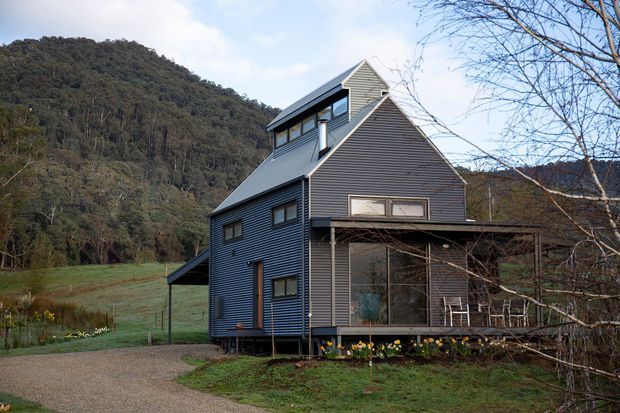 A multi-level contemporary house set in a wooded, rural mountain setting. The dark horizontal-clad building has steep gabled roof forms, extensive glazing, and a covered timber-framed veranda with outdoor seating. A gravel driveway, garden planting, and surrounding forested hills are visible.