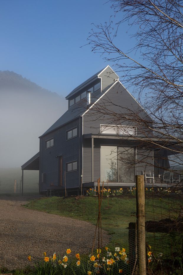 A tall, narrow rural house viewed from the front side in soft morning mist. The home has dark grey horizontal cladding, steep pitched gabled roofs, white trim, a projecting upper-level dormer-like volume, and a large ground-floor glazed opening. A gravel driveway, simple wire fencing, garden planting, and leafless trees surround the building, with hazy hills visible behind it.